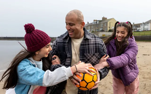 Father and two daughters laughing and playing with a football on a beach