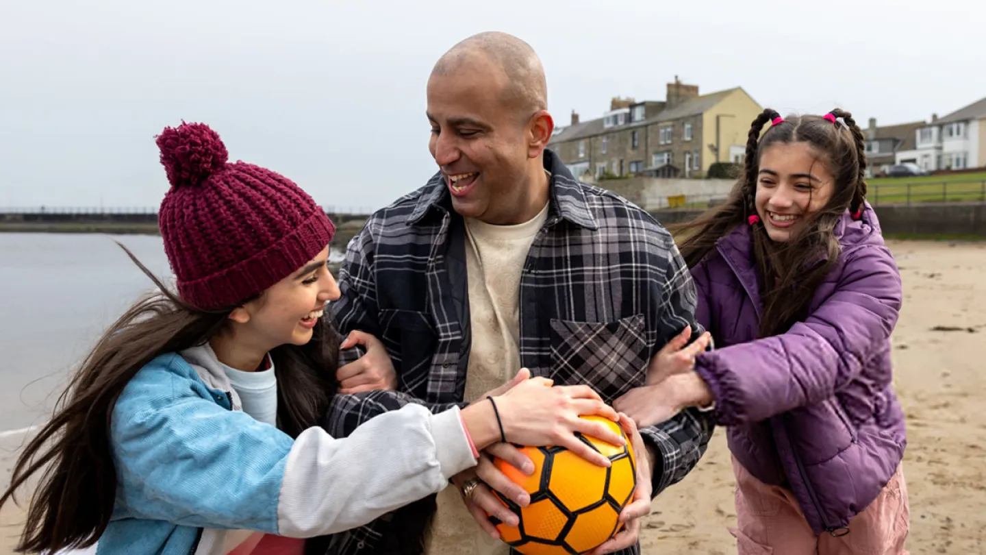 Father and two daughters laughing and playing with a football on a beach