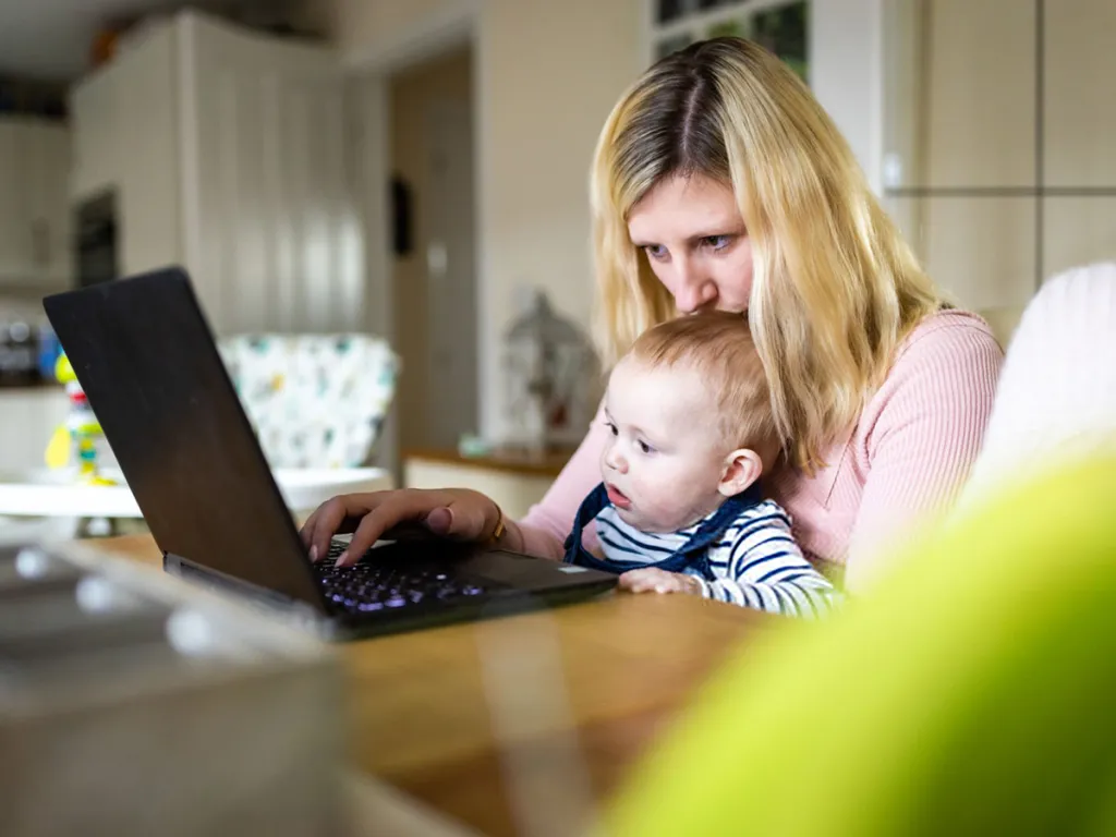 Woman working on a laptop at a kitchen table while holding a baby on her lap
