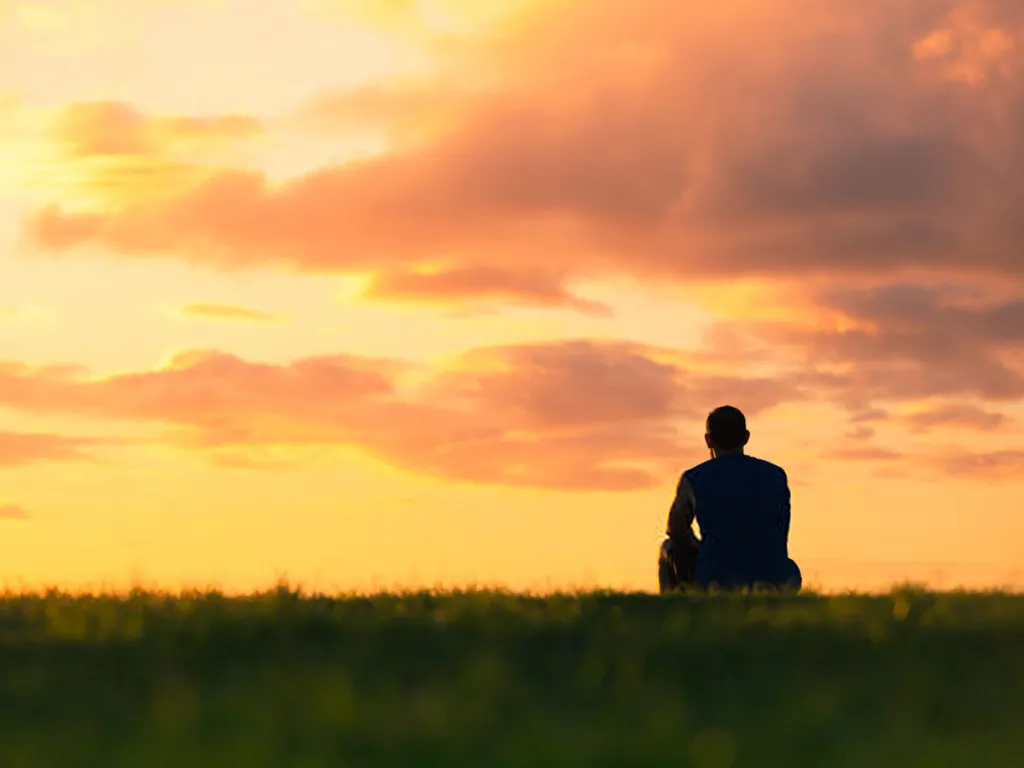 Person sitting alone on grass, looking out at a sunset sky